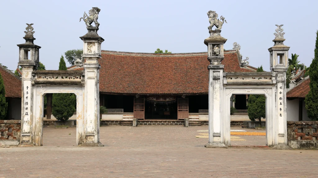 Derrière la porte La façade de la maison communale de Mông Phụ à Đường Lâm, avec son toit de tuiles, ses piliers et sa cour pavée.
