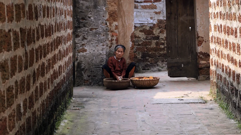 Derrière la porte Une femme accroupie devant deux paniers dans une ruelle de Đường Lâm, entre des murs en latérite et une porte en bois.