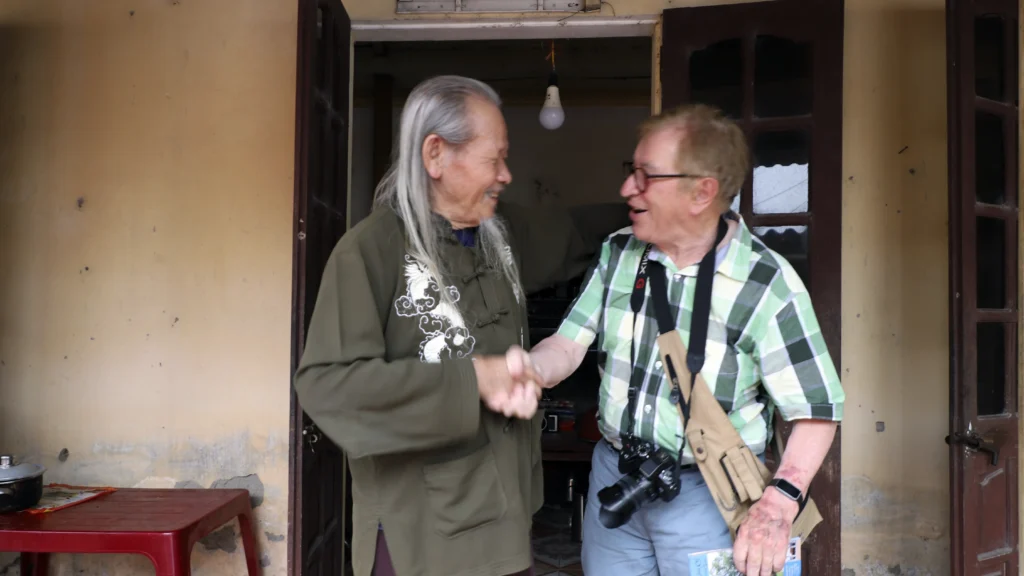 Derrière la porte Denis retrouve M. Đĩnh à Đường Lâm. Les deux hommes se serrent la main en souriant devant une porte ouverte.