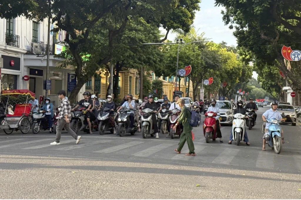 Pourquoi klaxonne-t-on autant au Vietnam ? Des conducteurs à moto attendent devant un passage piéton à un carrefour bordé d’arbres à Hanoi.