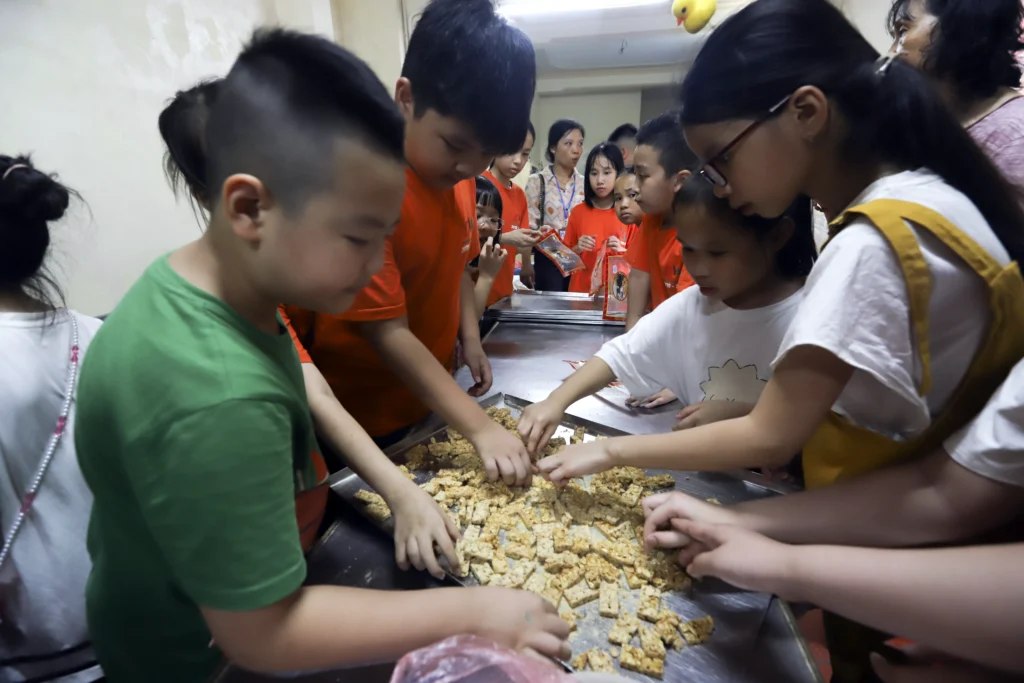 Derrière la porte Des enfants apprennent à fabriquer du kẹo lạc, une confiserie aux cacahuètes, autour d’une table à Đường Lâm.