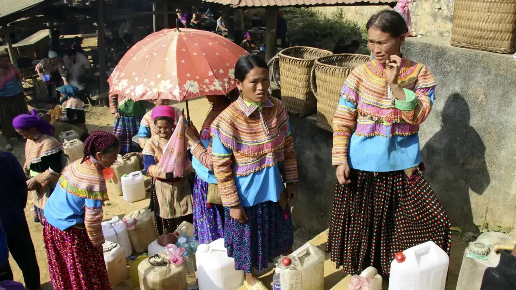 Des femmes H’Mông fleuries près de bidons d’alcool de maïs au marché de Bắc Hà.
