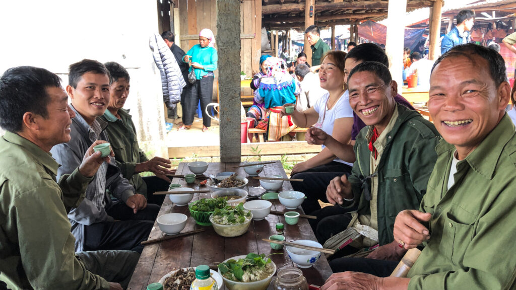 Des convives souriants autour d’une table garnie de bols et de petites tasses au marché de Cốc Ly.